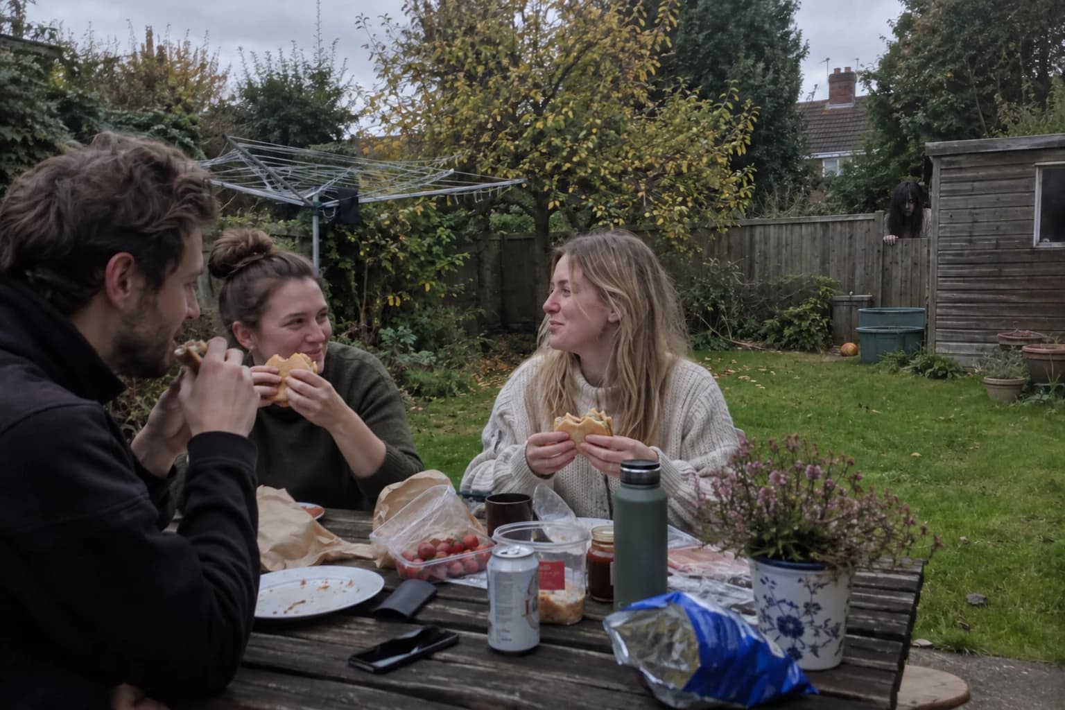 AI concept: friends at lunch while a long-haired figure watches from beside the garden shed