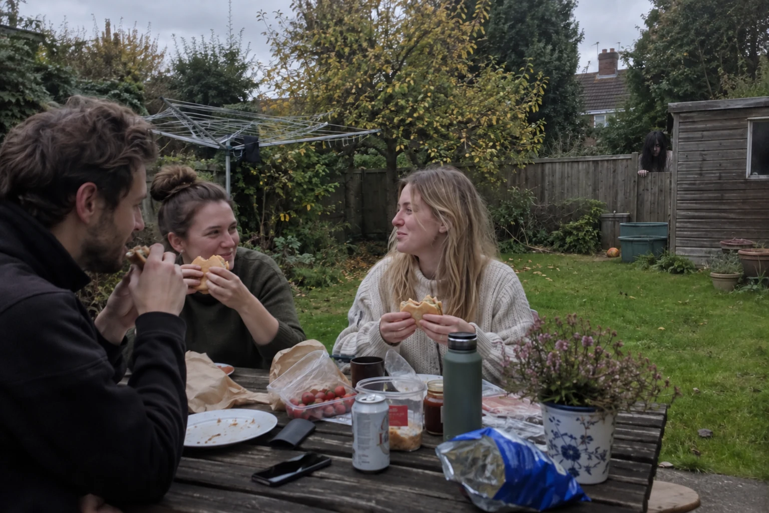 Friends at a garden lunch with a long-haired woman watching from the fence beside a shed.